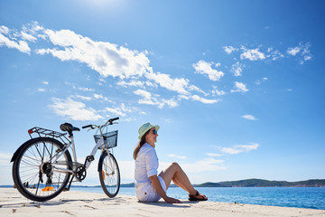 Fototapeta na wymiar Attractive smiling young woman in white closing, sunhat and sunglasses sitting at bicycle on stony sidewalk under clear blue sky on sparkling clear water background. Tourism and vacations concept.
