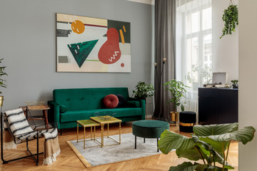 Luxury and modern home interior with design green sofa, commode, coffee tables, pouf and accessroies. A lot of plants in the room. Abstract painting. Stylish decor of living room with brown parquet. 