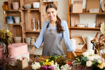 Waist up portrait of young female florist smiling at camera while arranging flowers and posing in flower shop