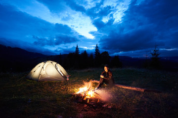 Fototapeta na wymiar Active woman tourist having a rest at night camping in the mountains, sitting near burning campfire and illuminated tourist tent under beautiful evening cloudy sky. Tourism, outdoor activity concept