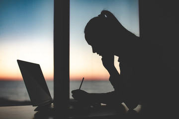 Silhouette of a business woman working late at a laptop against the window at sunset. Night and evening online working