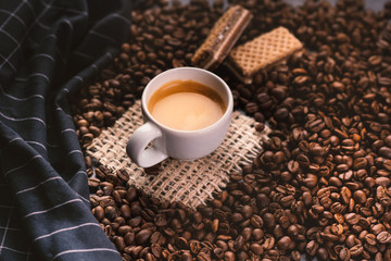 Cup of coffee in the middle of coffee beans with biscuits and tablecloth. Grained product. Hot drink. Close up. Harvesting. Natural background. Energy. Fresh espresso coffee