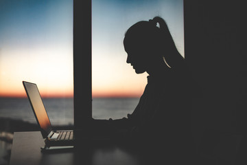 Silhouette of a business woman working late at a laptop against the window at sunset. Night and evening online working