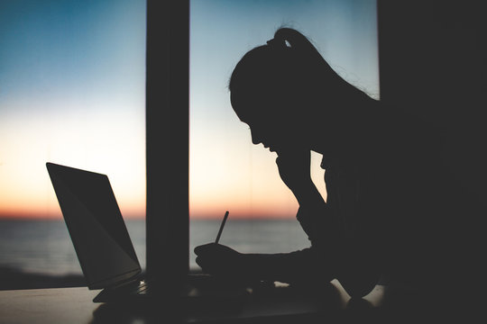 Silhouette Of A Business Woman Working Late At A Laptop Against The Window At Sunset. Night And Evening Online Working
