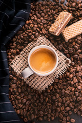 Cup of coffee in the middle of coffee beans with biscuits and tablecloth. Grained product. Hot drink. Close up. Harvesting. Natural background. Energy. Fresh espresso coffee