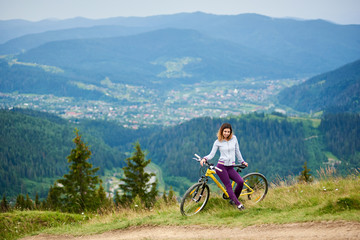Fototapeta premium Happy female cyclist resting on yellow bicycle in the mountains on cloudy evening. Mountains, forests and small city on the blurred background. Outdoor sport activity, lifestyle concept