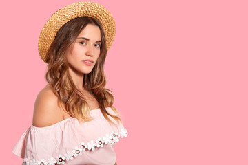 Horizontal shot of pleasant looking young woman stands bare shoulders, wears summer hat and fashionable blouse, thinks about somehting, isolated over pink background with free space for text