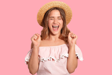 Isolated shot of overjoyed young woman clenches fists with happiness, closes eyes from pleasure, exclaims with triumph, wears fashionable clothes, isolated over pink background. Success concept