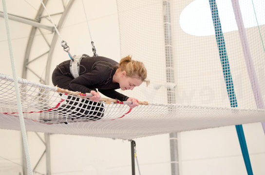 An adult female lands on a net, preparing to dismount at a on a flying trapeze school at an indoor gym. The woman is an amateur trapeze artist.