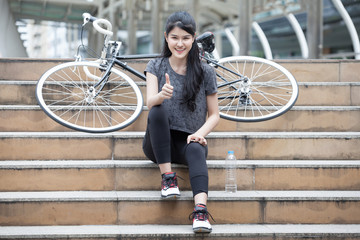 asian young sport woman in sportswear sitting on the stairs with bicycle and showing thumbs up ....