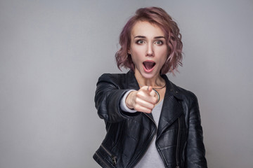 Portrait of surprised beautiful girl with short hairstyle and makeup in black leather jacket standing, pointing and looking at camera with amazed face. indoor studio shot, isolated on grey background.