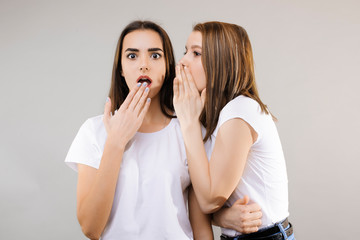 Two beautiful female dressed in white shirts gossiping. Two cute girls posing at the camera while one is whispering a secret on another's ear while another opens hers mouth amazed .