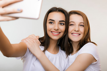 Two amazing young woman doing a selfie smiling and hugging in front of a white wall. Beautiful girlfriends having fun while making a selfie with a white smartphone against white background.