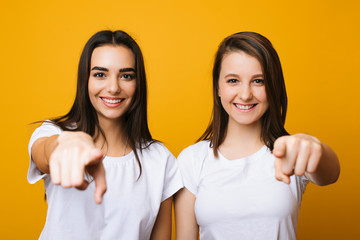 Portrait of two lovely young girls looking looking into camera and pointing at the camera with their finger smiling against yellow wall. Two girlfriends pointing on you with her fingers.