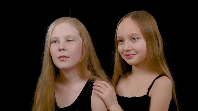 Two cute little girls friends looking sideward. Close up view happy beautiful teenage models girls with long hair, standing together and looking aside, isolated on black background