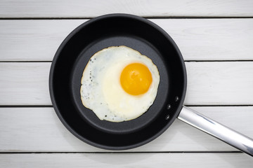 Top view of a sunny side up fried egg in a black pan on wooden white table. 