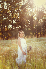 Fototapeta na wymiar Beautiful and happy pregnant woman in a white dress on the nature in the summer, around the trees and flowers.