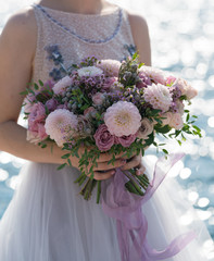 the bride holds a pink and lilac wedding bouquet in her arms against the background of the sea