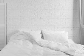 Large comfortable bed with pillows and blanket near white brick wall indoors. Stylish interior