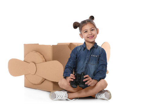Cute little girl playing with binoculars and cardboard airplane on white background