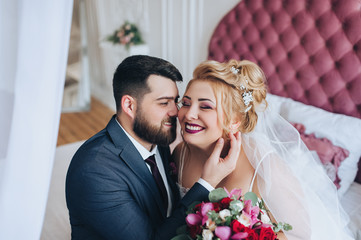 Stylish bearded groom and beautiful blonde bride hugging on a bed in a white studio. Portrait of newlyweds in love in the interior. Wedding photography.