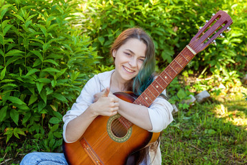 Young hipster woman sitting in grass and playing guitar on park or garden background. Teen girl...
