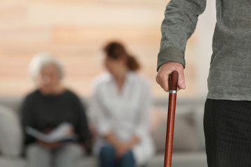 Elderly man holding walking cane and blurred caregiver with senior woman on background, focus on hand. Space for text