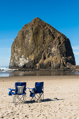 Fototapeta na wymiar Blue beach chairs put in front of the huge Haystack rock in Cannon Beach, Oregon, USA.
