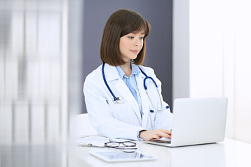 Doctor typing on laptop computer while sitting at the white table in hospital office. Physician at work. Medicine and healthcare concept