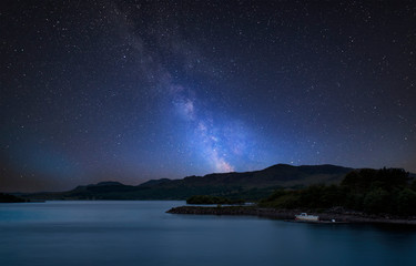 Stunning vibrant Milky Way composite image over landscape of calm lake with boat on shore