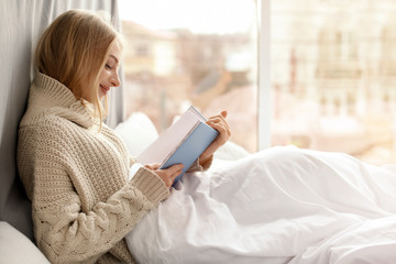 Beautiful young woman in knitted sweater sitting and reading book near window at home. Winter atmosphere