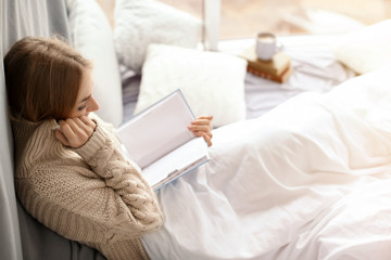 Beautiful young woman in knitted sweater sitting and reading book near window at home. Winter...