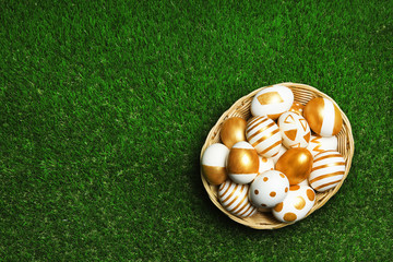 Naklejka premium Wicker basket of traditional Easter eggs decorated with golden paint on green lawn, top view. Space for text