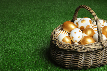 Wicker basket of traditional Easter eggs decorated with golden paint on green lawn, space for text