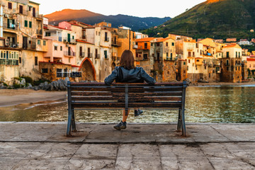 Young woman sitting on the bench and enjoying view of coastal village Cefalu at sunrise, Sicily,...