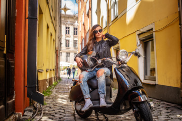 Plakat Girl in sunglasses wearing a leather jacket and ripped jeans sitting on a black classic scooter on an old narrow street.