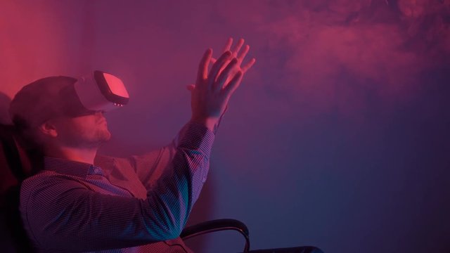 Cute guy in shirt using VR headset interacts with virtual reality. He sits in a smoky dark room with red and blue lighting and waves his hands