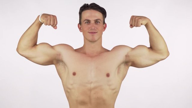Young cheerful bodybuilder flexing his biceps, smiling to the camera. Handsome shirtless sportsman showing off his strong muscular body. Happiness, strength, gym concept