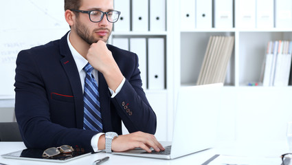 Businessman using laptop computer while sitting at the desk in office. Focus at cheerful smiling bearded man wearing glasses. Work place and success concept