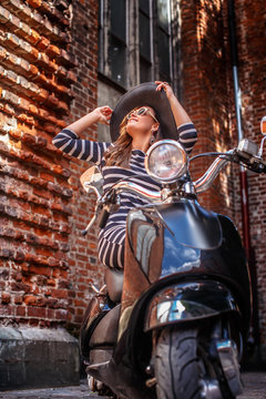Fototapeta Woman wearing dress and hat sitting on a black classic Italian scooter on an old street in Europe.