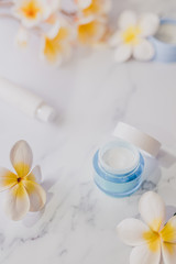 group of skincare products including moisturiser scrub and hand cream pots on marble table with exotic frangipani flowers