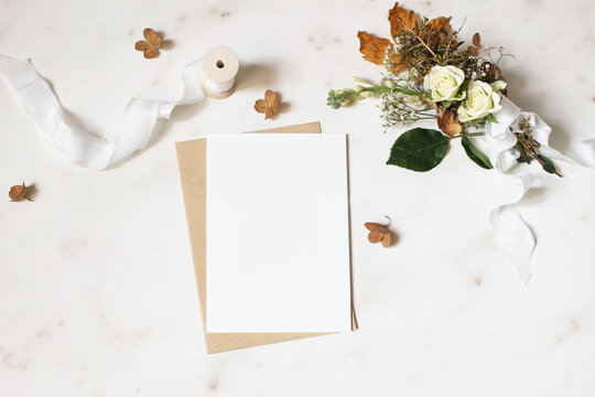 Feminine winter wedding, birthday stationery mock-up scene. Blank greeting card, envelope. Dry hydrangea, white roses and gypsophila flowers bouquet. Marble stone table background. Flat lay, top view.