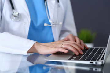 Doctor typing on laptop computer while sitting at the glass desk in hospital office. Physician at work. Medicine and healthcare concept