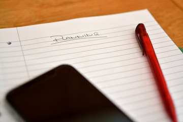 Closeup of lined paper with 'planning' written on the top. Red pen and black tablet / phone also in...