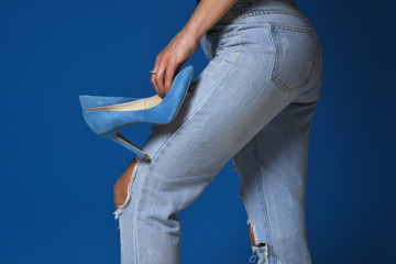 Clothes designer. Woman holds blue high heels on legs. Young girl prepares for walk. Graceful girl in blue torm jeans and blue heels on grey background. Fit legs and buttoicks of girl. Before the date