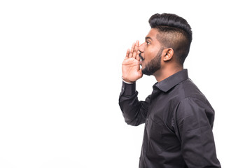 Portrait of Indian guy shouting loud, mouth open and hand cupped beside. Asian man standing on plain background with shadow and copy space.