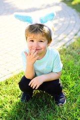 Funny kid. Cute boy with shy smile sits on grass and holds his hand near face. Little child with...