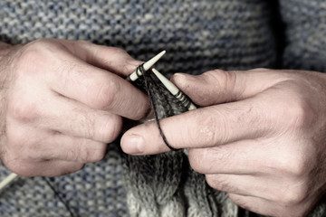 European man 40-50 years old knits a scarf. A man in a knitted sweater on the background of shelves with yarn and knitted products. Knitting like hobbies handmade. Hands knitting men close-up.
