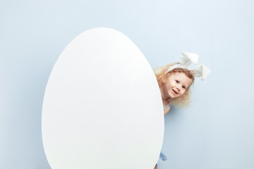 Little curly girl with bunny ears on her head hiding behind a large white egg against a blue wall....