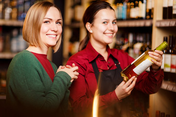 Photo of two smiling women with bottle of wine in store on background of shelves